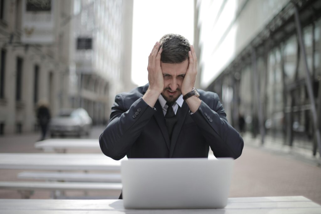 A businessman in a suit looks stressed, sitting outdoors with a laptop in a city environment.